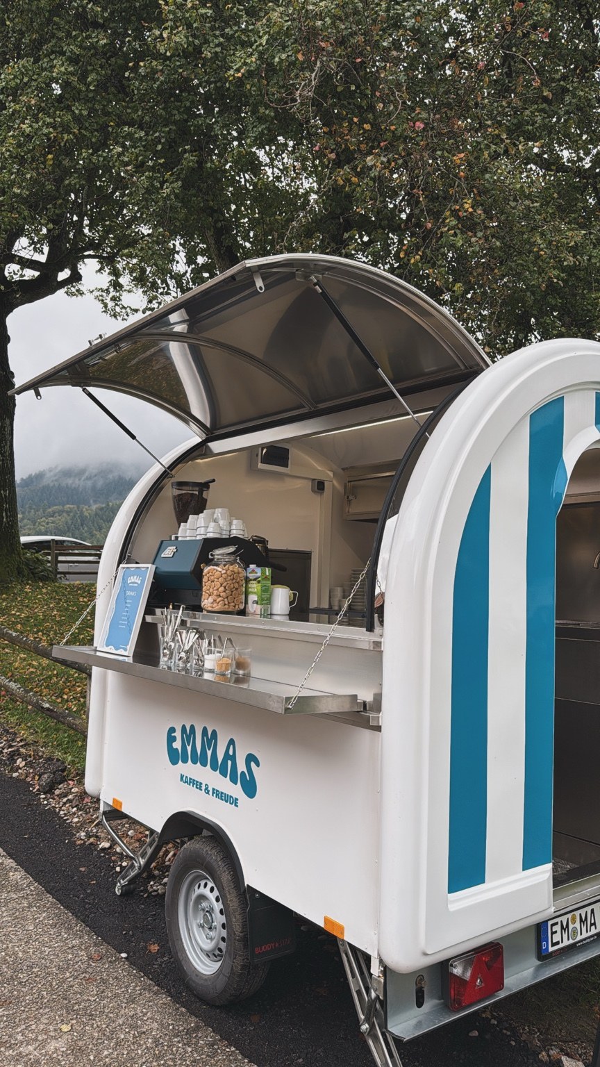 Emmas blau-weißer Kaffeewagen mit geöffneter Theke unter einem Baum